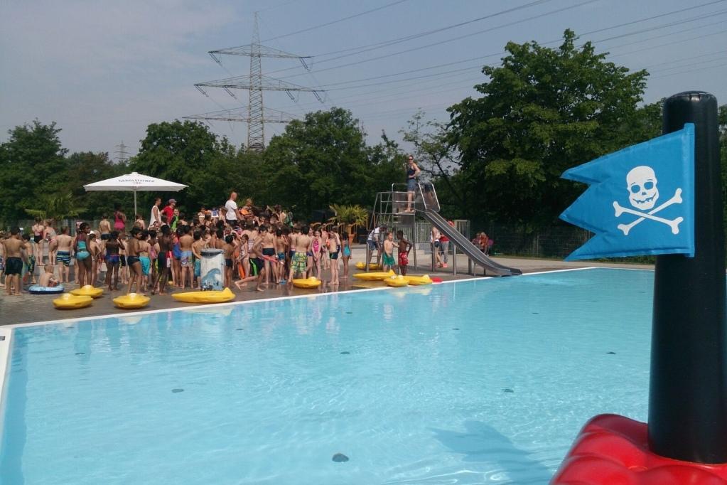 Blick auf einen Freibadbereich mit vielen Kindern, Rutsche und gelben Schwimmfl&uuml;gen unter blauem Himmel.