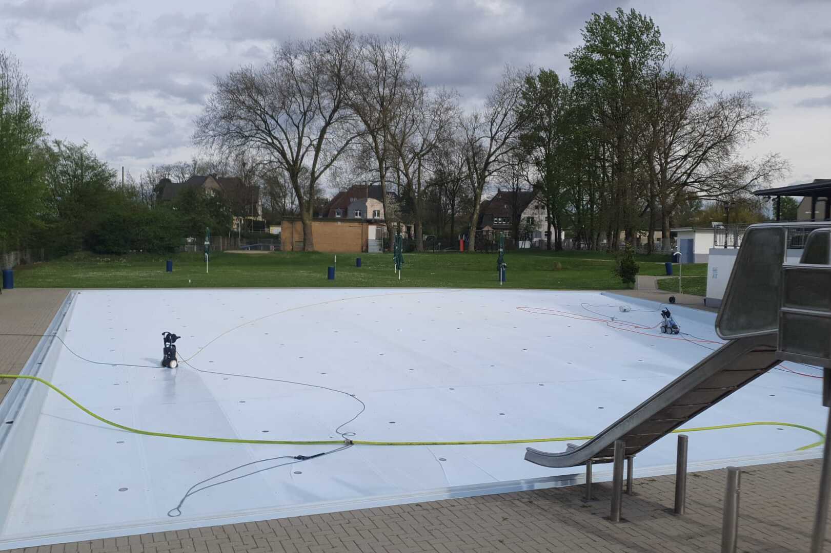 Leeres Schwimmbecken mit Wasserrutschen und Gartenanlage, umgeben von B&auml;umen, an einem bew&ouml;lkten Tag.