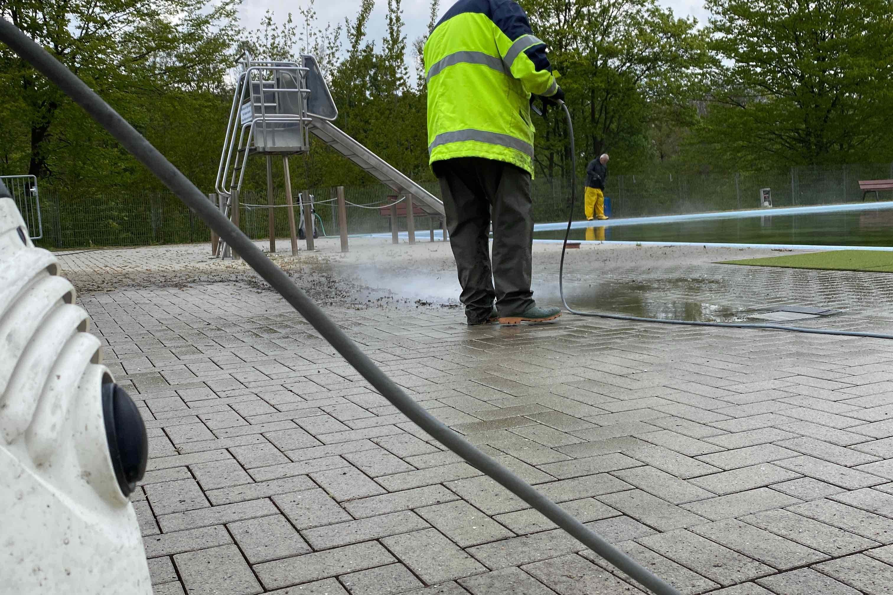 Mann in reflektierender Jacke reinigt mit Hochdruckreiniger eine gepflasterte Fl&auml;che neben einem Spielplatz.