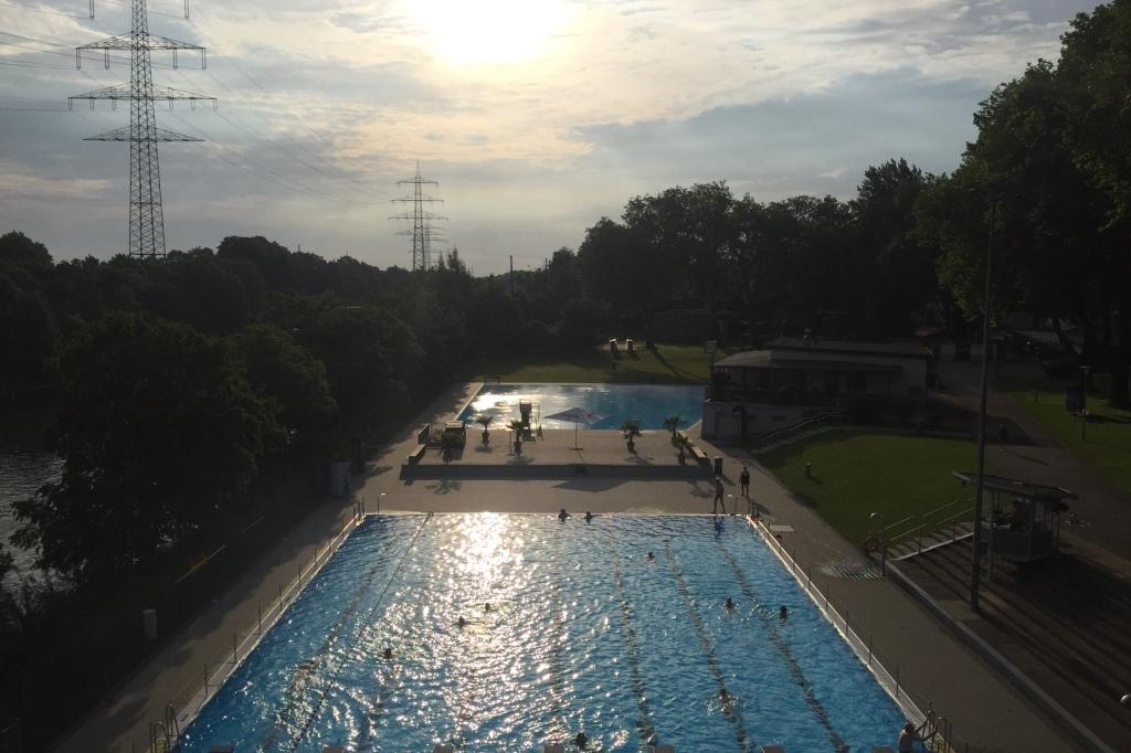 Sonnenuntergang &uuml;ber einem Schwimmbad mit blauem Wasser und B&auml;umen, mit Schwimmern und einem Freizeitbereich im Hintergrund.