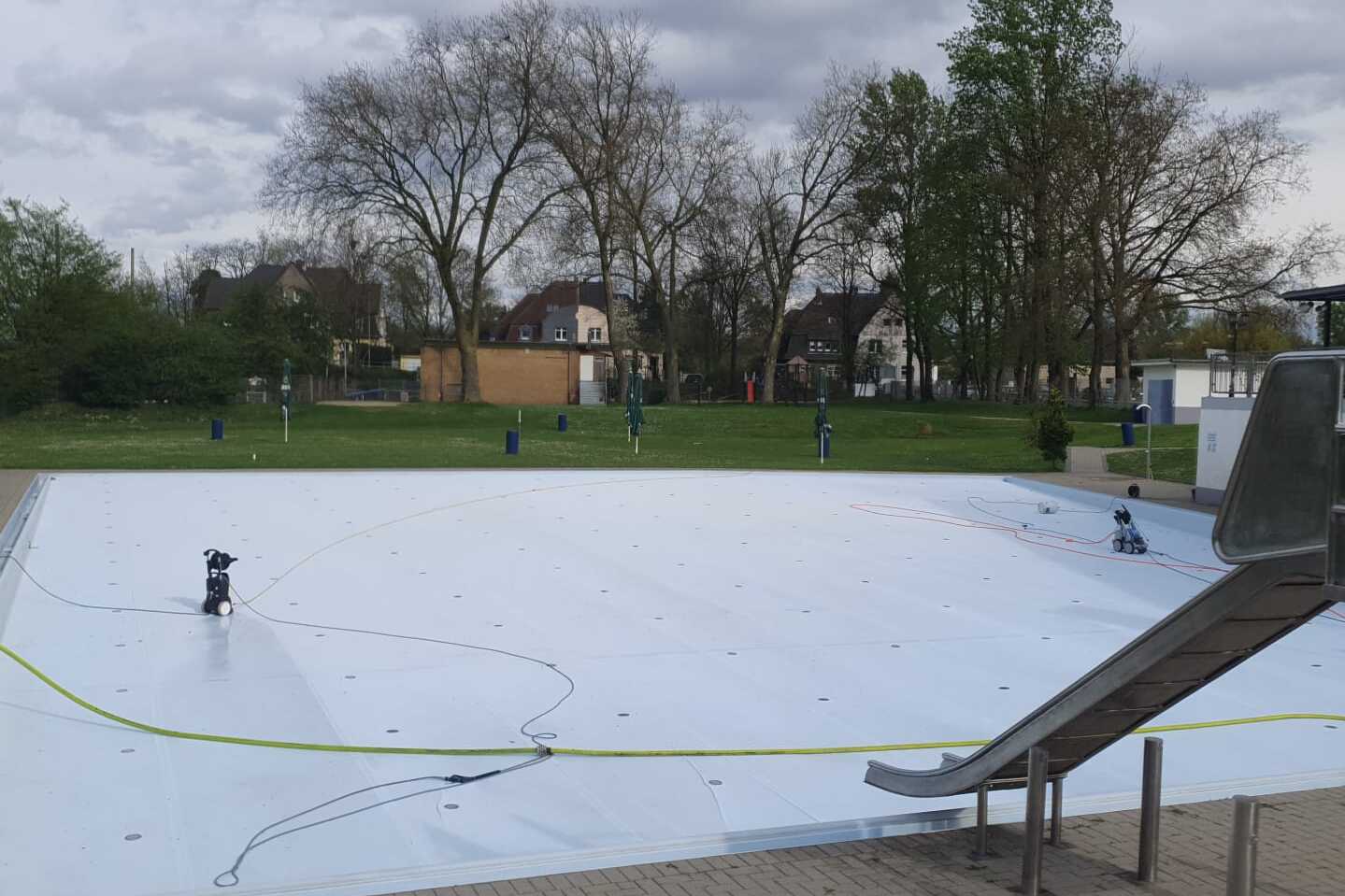 Leeres Schwimmbecken mit Wasserrutschen und Gartenanlage, umgeben von B&auml;umen, an einem bew&ouml;lkten Tag.