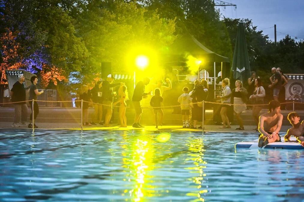 Abendliche Stimmung beim Nachtschwimmen im Freibad &bdquo;Hesse" Dellwig: G&auml;ste tanzen und feiern am Beckenrand unter gelbem und blauem B&uuml;hnenlicht, w&auml;hrend im Vordergrund zwei Jugendliche auf einer Schwimminsel im beleuchteten Wasser