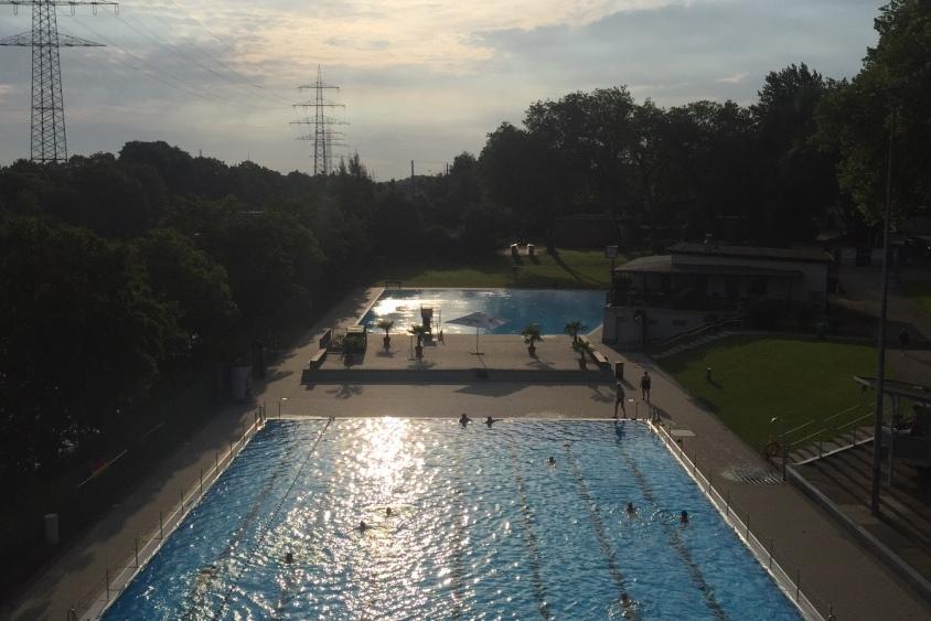 Sonnenuntergang &uuml;ber einem Schwimmbad mit blauem Wasser und B&auml;umen, mit Schwimmern und einem Freizeitbereich im Hintergrund.
