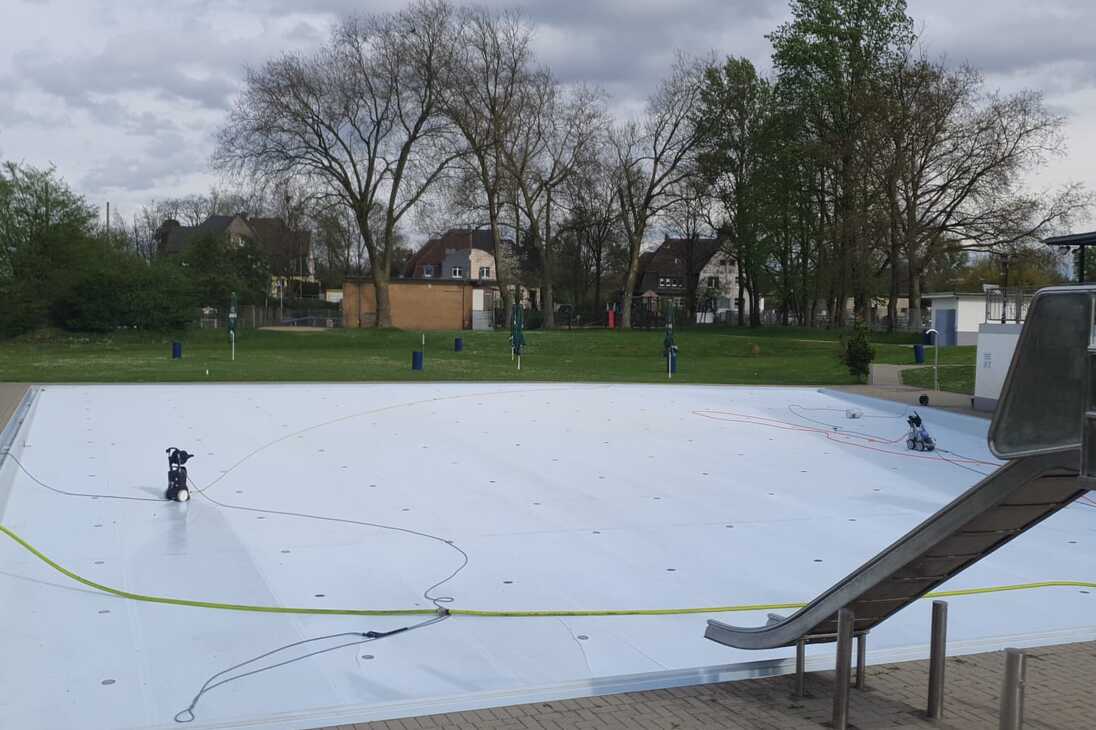 Leeres Schwimmbecken mit Wasserrutschen und Gartenanlage, umgeben von B&auml;umen, an einem bew&ouml;lkten Tag.