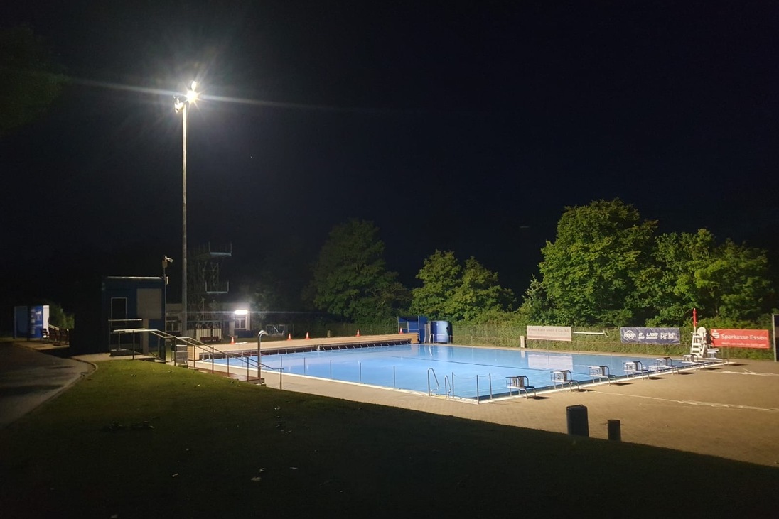 Beleuchtetes Schwimmbad mit blauer Wasserfl&auml;che und umliegendem Gr&uuml;n bei Nacht.