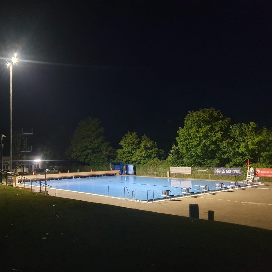 Beleuchtetes Schwimmbad mit blauer Wasserfl&auml;che und umliegendem Gr&uuml;n bei Nacht.