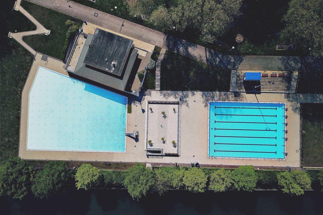 Luftaufnahme eines Schwimmbades mit einem gro&szlig;en und einem Sportbecken, umgeben von B&auml;umen und Wegen.
