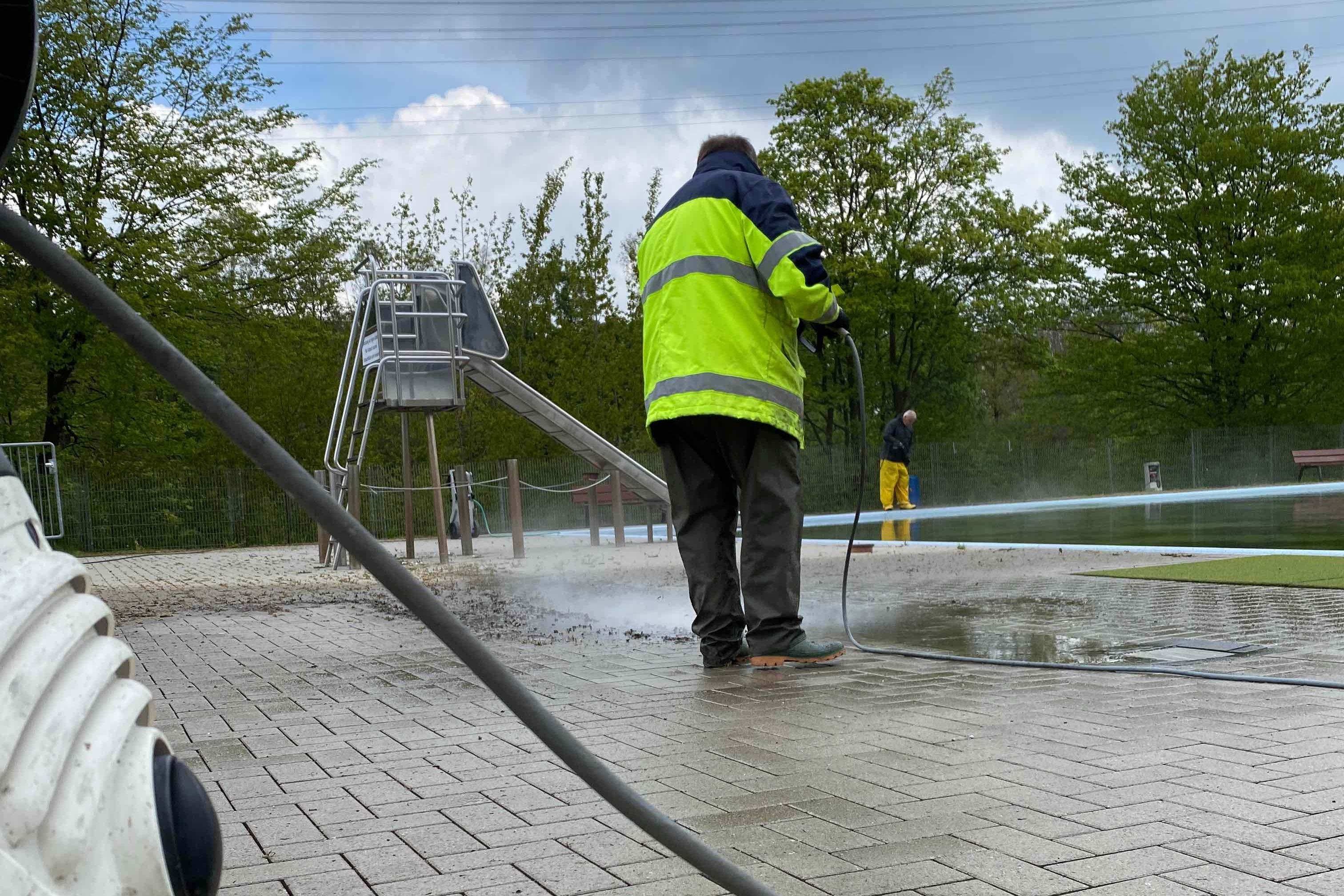 Mann in reflektierender Jacke reinigt mit Hochdruckreiniger eine gepflasterte Fläche neben einem Spielplatz.
