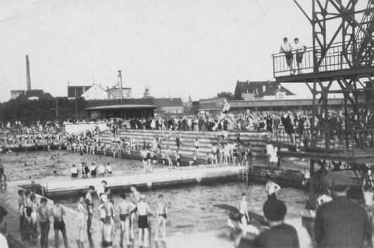 Blick auf ein belebtes Freibad mit Schwimmern und Zuschauern, im Hintergrund Trib&uuml;nen und einen Sprungturm.