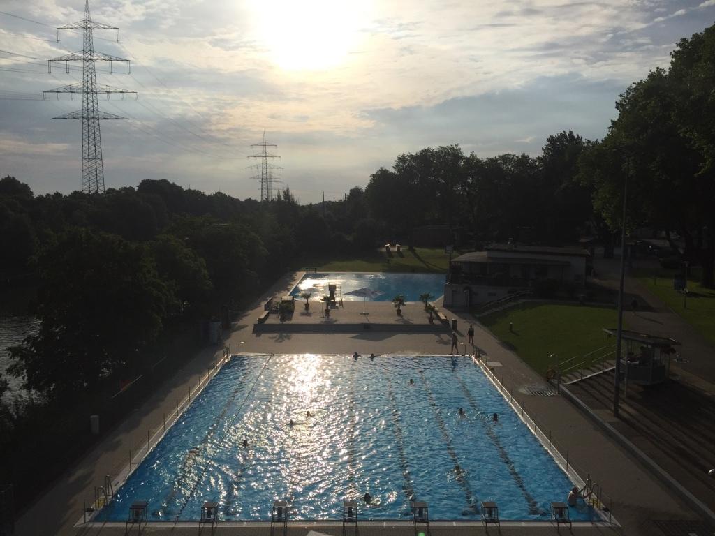 Sonnenuntergang &uuml;ber einem Schwimmbad mit blauem Wasser und B&auml;umen, mit Schwimmern und einem Freizeitbereich im Hintergrund.