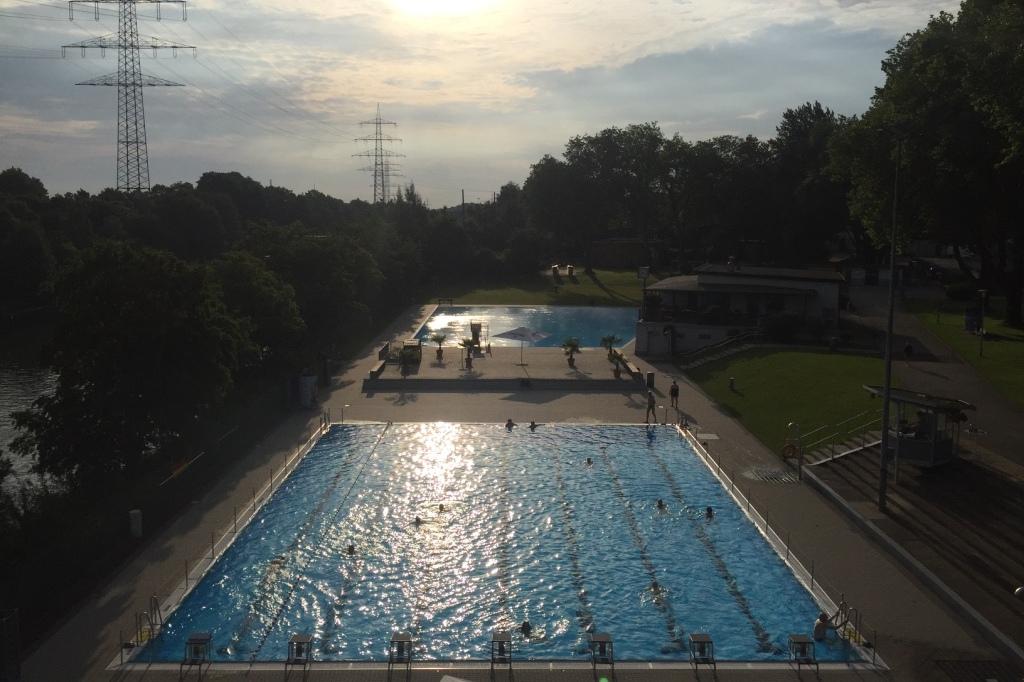 Sonnenuntergang &uuml;ber einem Schwimmbad mit blauem Wasser und B&auml;umen, mit Schwimmern und einem Freizeitbereich im Hintergrund.