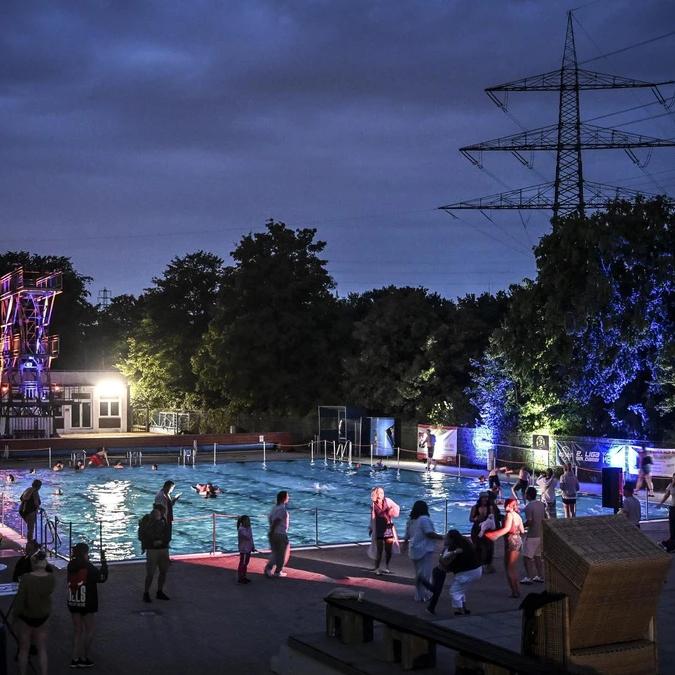 Nachtschwimmen 2026 Blick über das illuminierte Freibad Dellwig beim Nachtschwimmen: Gäste im Wasser und am Beckenrand, im Hintergrund der bunt angestrahlte Sprungturm.