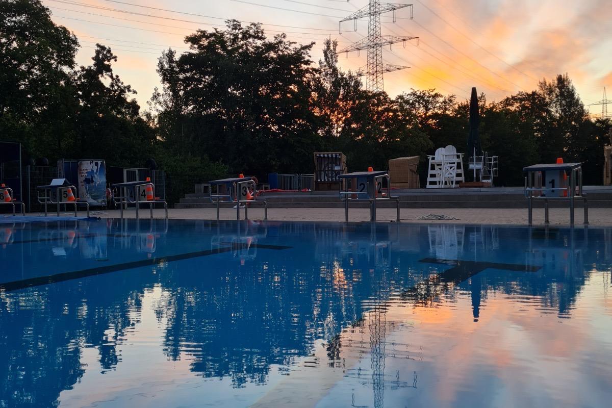 Schwimmerbecken des Freibads &bdquo;Hesse" Dellwig am Abend: Das ruhige, blaue Wasser spiegelt den orange-rosa gef&auml;rbten Sonnenuntergangshimmel und die umliegenden B&auml;ume wider. Am Beckenrand stehen nummerierte Startbl&ouml;cke, im Hintergru