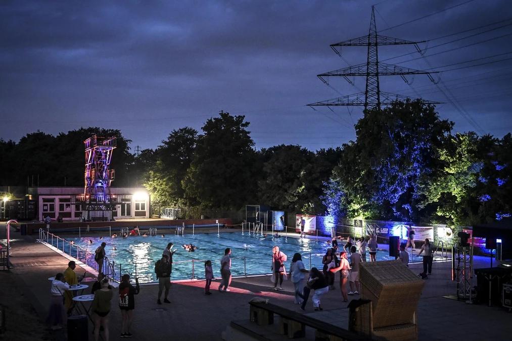 Nachtschwimmen 2026 Blick über das illuminierte Freibad Dellwig beim Nachtschwimmen: Gäste im Wasser und am Beckenrand, im Hintergrund der bunt angestrahlte Sprungturm.