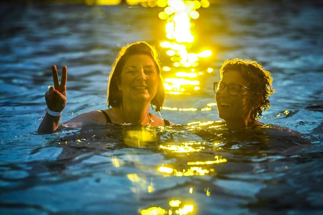 Zwei lachende Schwimmerinnen im abendlich beleuchteten Becken beim Nachtschwimmen, auf dem Wasser spiegelt sich goldenes Licht.