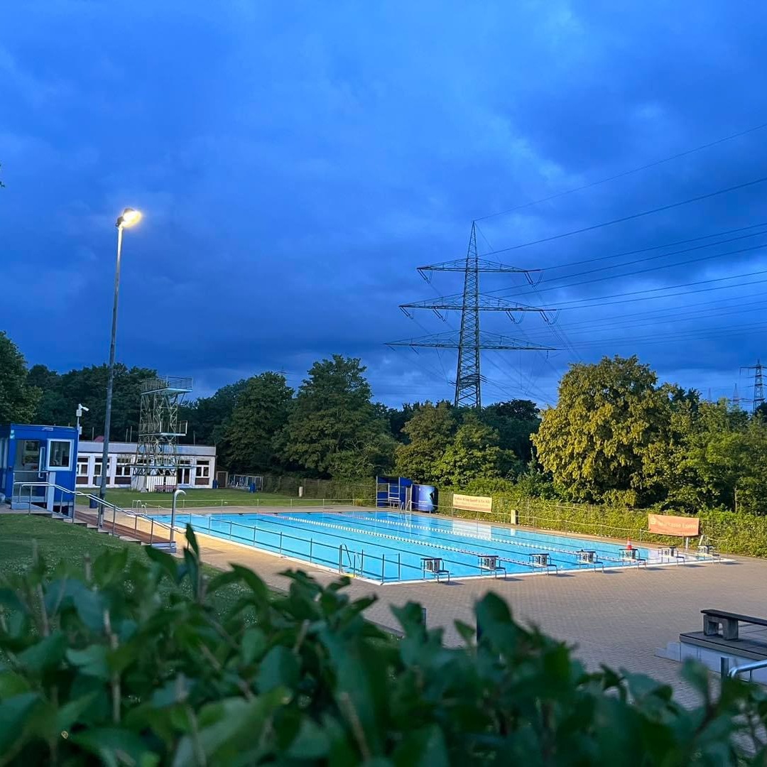 Blick auf ein beleuchtetes Schwimmbad mit gr&uuml;ner Umgebung und dunklem Himmel, umgeben von B&auml;umen und Strommasten.