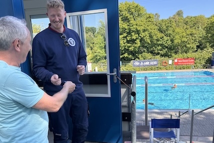 Zwei M&auml;nner stehen am Eingang eines Schwimmbadgeb&auml;udes, im Hintergrund ist ein Schwimmbecken und B&auml;ume sichtbar.