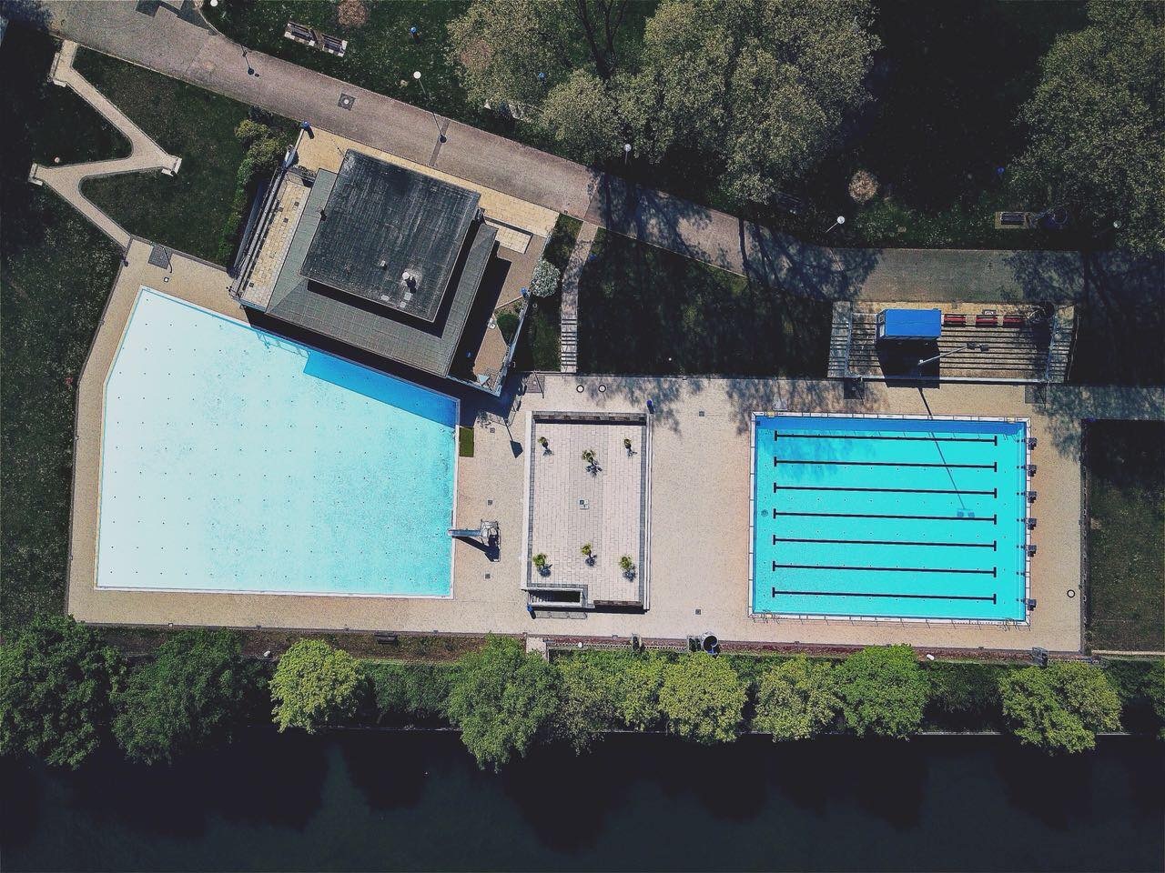 Luftaufnahme eines Schwimmbades mit einem gro&szlig;en und einem Sportbecken, umgeben von B&auml;umen und Wegen.
