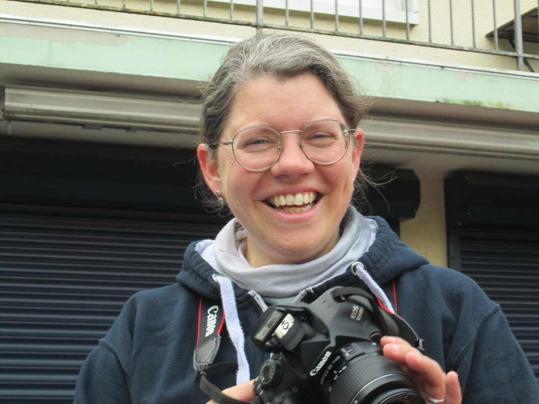 L&auml;chelnde Person mit Brille h&auml;lt eine Kamera und steht vor einer grauen Wand.