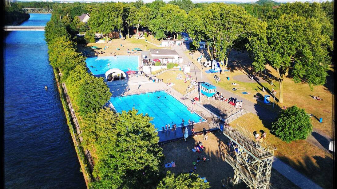 Luftaufnahme eines Schwimmbads mit zwei Becken und Liegewiese, umgeben von B&auml;umen und einem Fluss im Hintergrund.