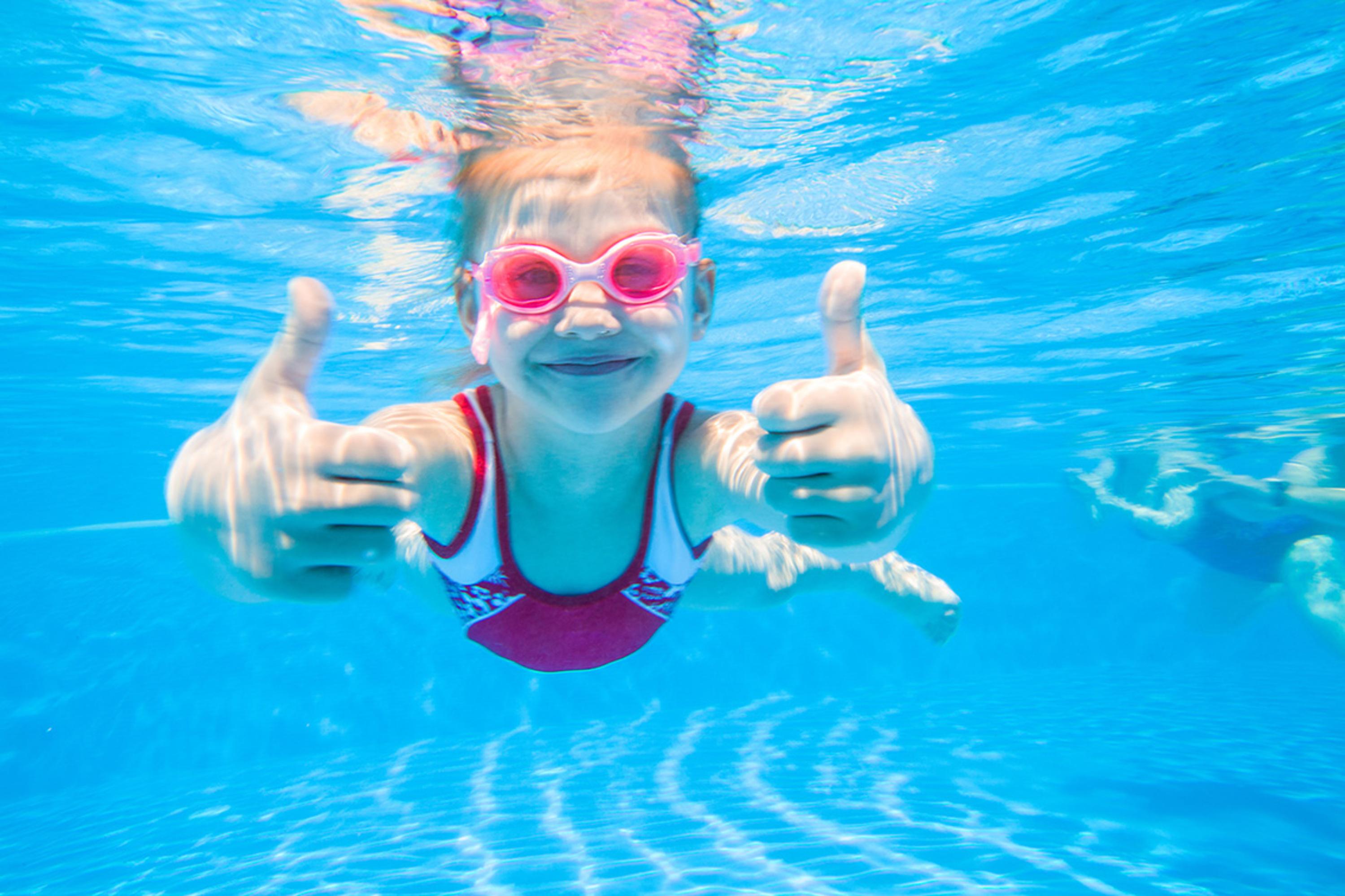 Lächelndes Mädchen mit Schwimmbrille zeigt Daumen hoch unter Wasser in einem blauen Schwimmbecken.