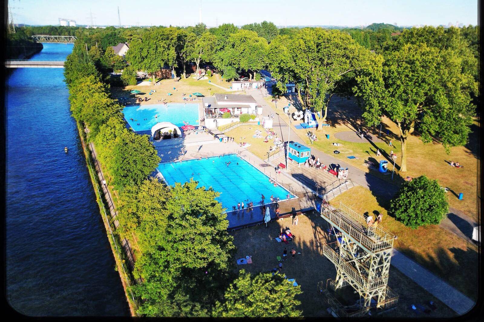 Luftaufnahme eines Schwimmbads mit zwei Becken und Liegewiese, umgeben von B&auml;umen und einem Fluss im Hintergrund.