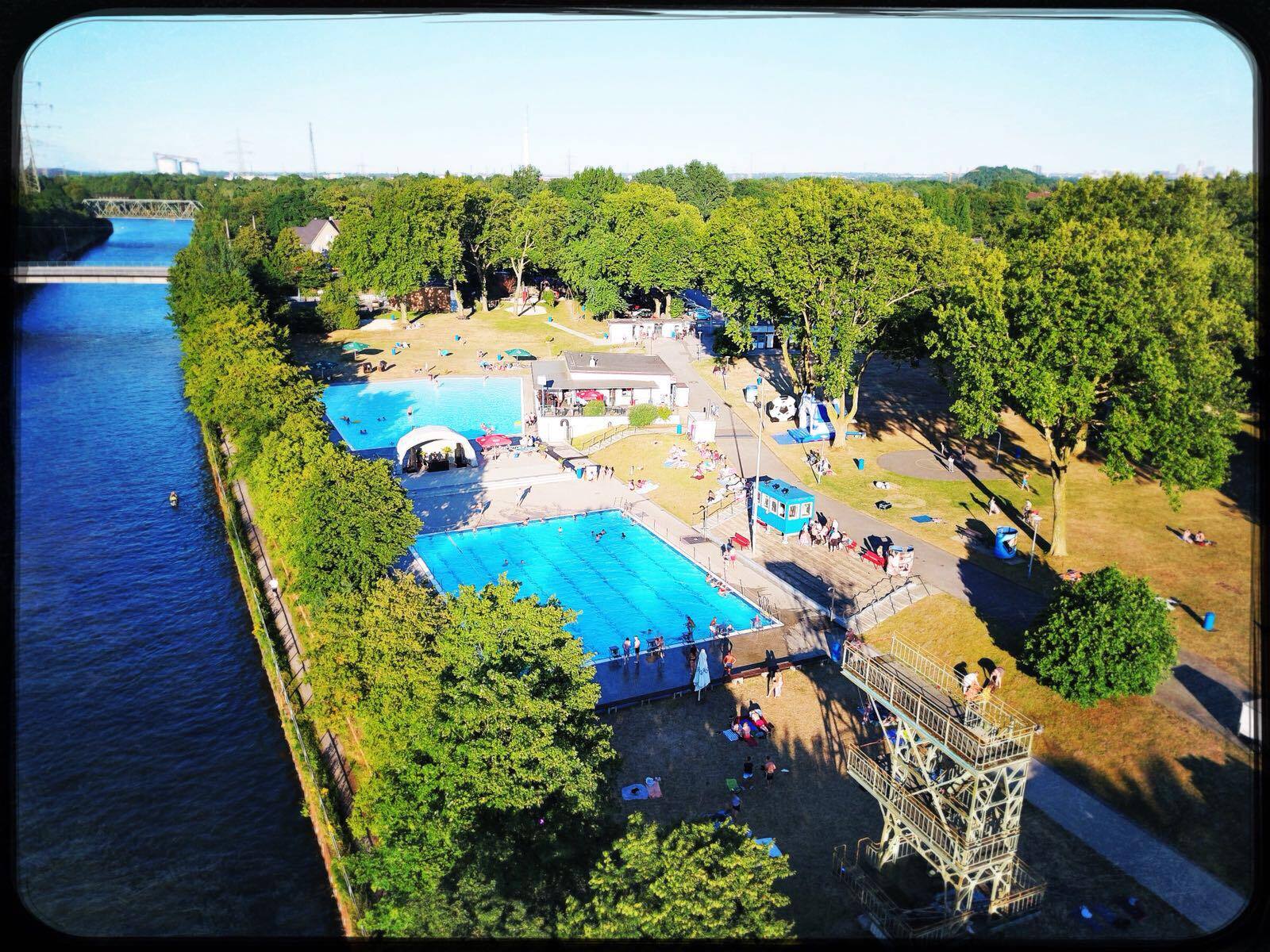 Luftaufnahme eines Schwimmbads mit zwei Becken und Liegewiese, umgeben von B&auml;umen und einem Fluss im Hintergrund.