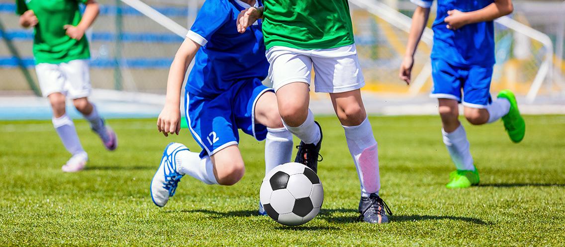 Spielende Kinder in blauen und gr&uuml;nen Trikots dribbeln einen Fu&szlig;ball auf einem Rasenplatz.
