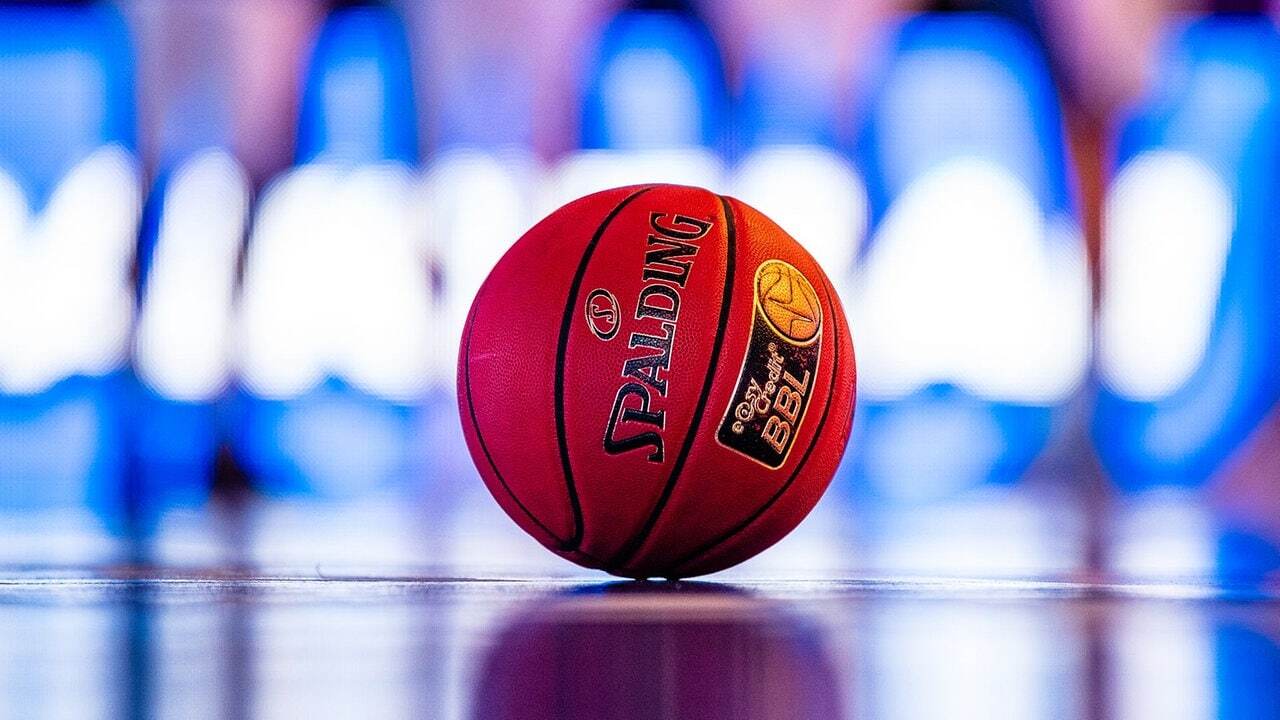 Nahaufnahme eines rot-orangenen Basketballs auf einem gl&auml;nzenden Spielfeld, verschwommene Spieler im Hintergrund.