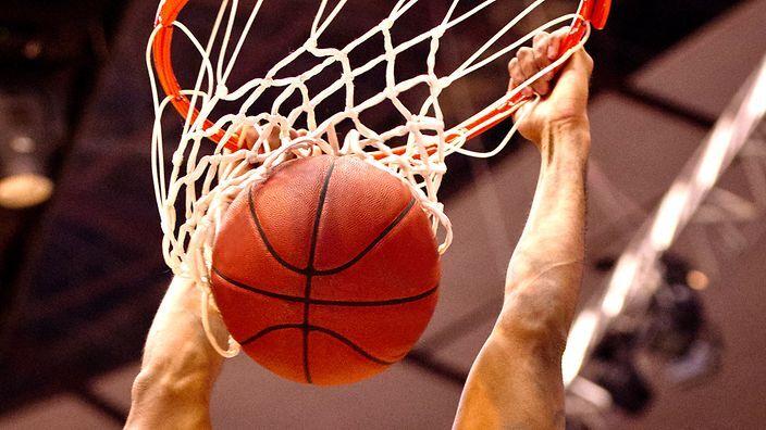 Ball landet im Basketballkorb, w&auml;hrend eine Hand den Netzrahmen h&auml;lt, mit verschwommenem Hintergrund.