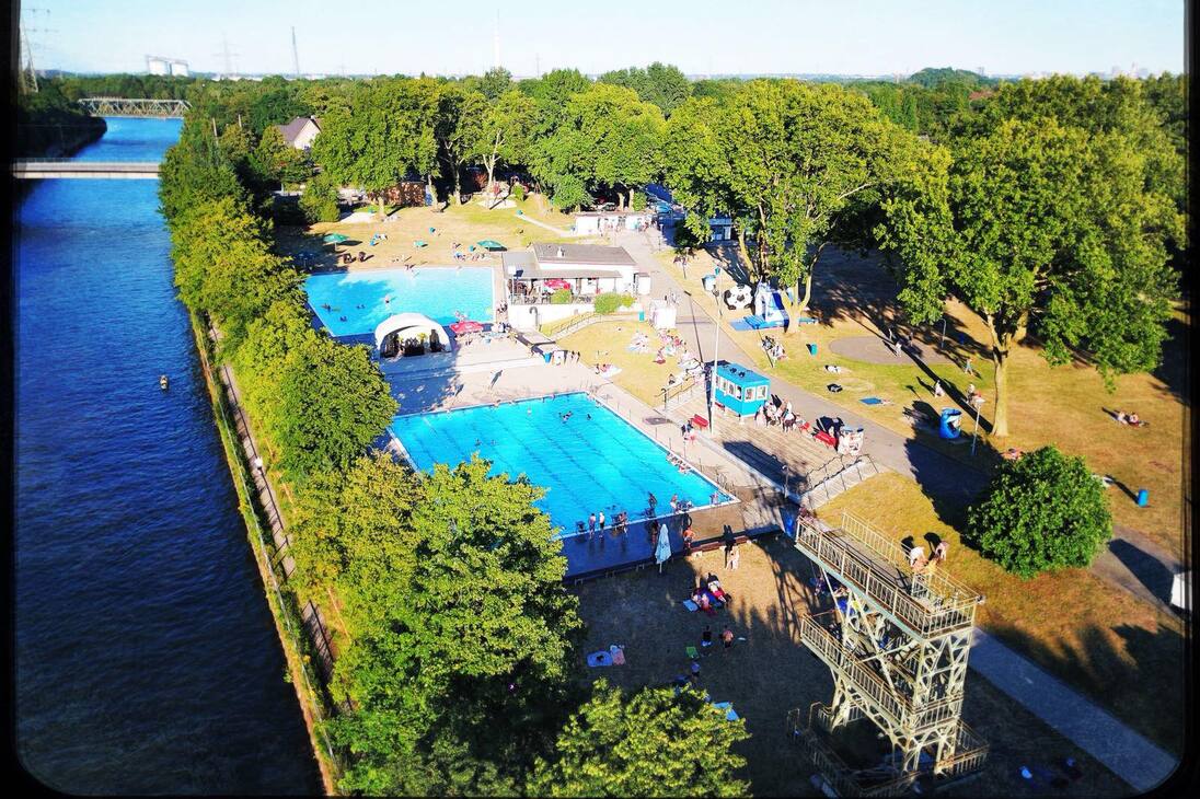 Luftaufnahme eines Schwimmbads mit zwei Becken und Liegewiese, umgeben von B&auml;umen und einem Fluss im Hintergrund.