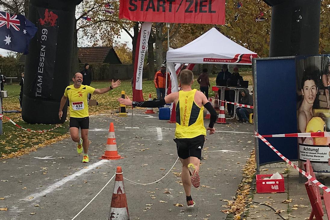 Zwei L&auml;ufer erreichen unter einem Bogen mit der Aufschrift &bdquo;START/ZIEL&ldquo; das Ziel eines Wettkampfs auf einer herbstlichen Strecke.