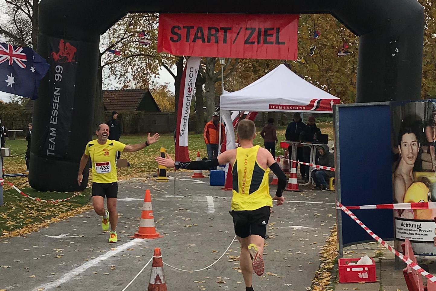 Zwei L&auml;ufer erreichen unter einem Bogen mit der Aufschrift &bdquo;START/ZIEL&ldquo; das Ziel eines Wettkampfs auf einer herbstlichen Strecke.