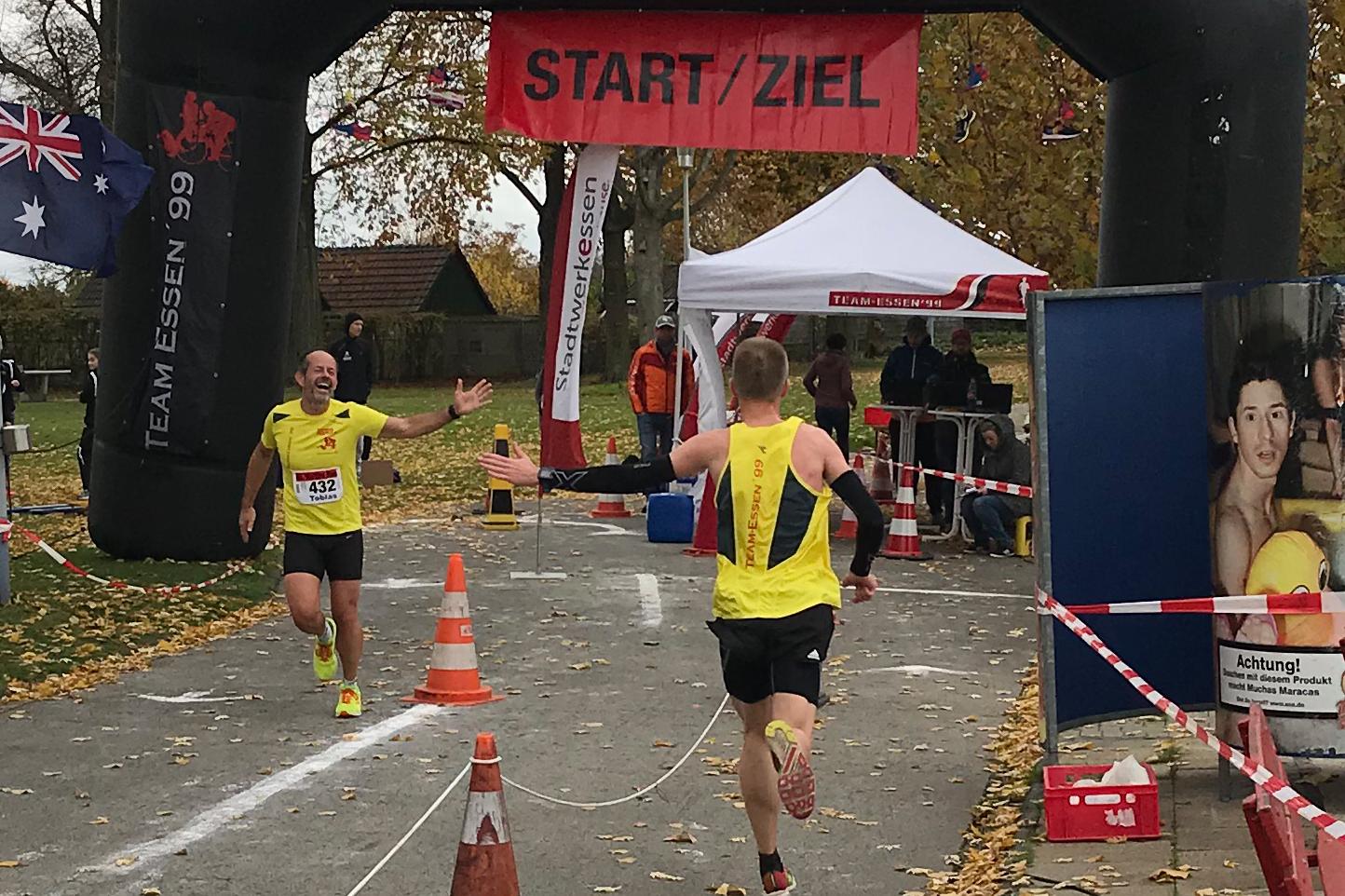 Zwei Läufer erreichen unter einem Bogen mit der Aufschrift „START/ZIEL“ das Ziel eines Wettkampfs auf einer herbstlichen Strecke.