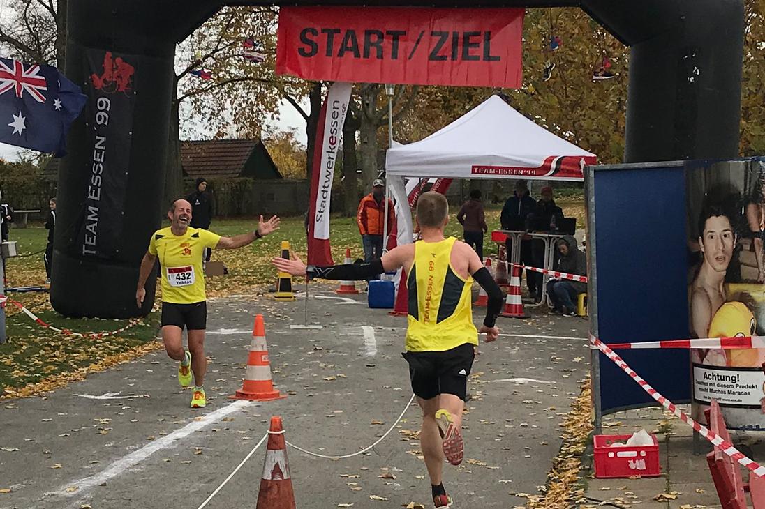 Zwei L&auml;ufer erreichen unter einem Bogen mit der Aufschrift &bdquo;START/ZIEL&ldquo; das Ziel eines Wettkampfs auf einer herbstlichen Strecke.