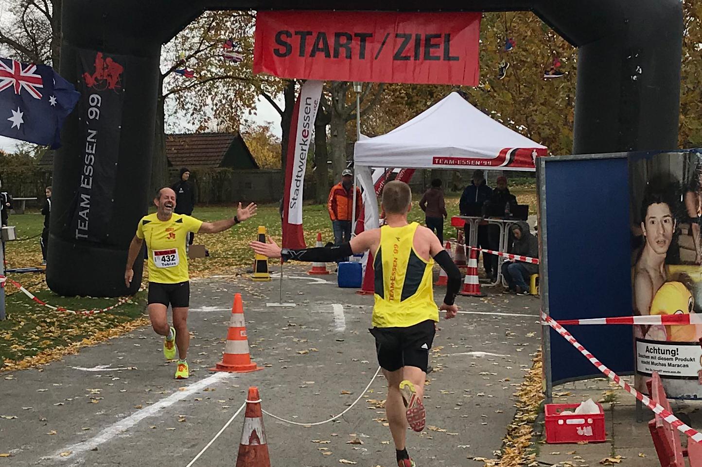 Zwei Läufer erreichen unter einem Bogen mit der Aufschrift „START/ZIEL“ das Ziel eines Wettkampfs auf einer herbstlichen Strecke.