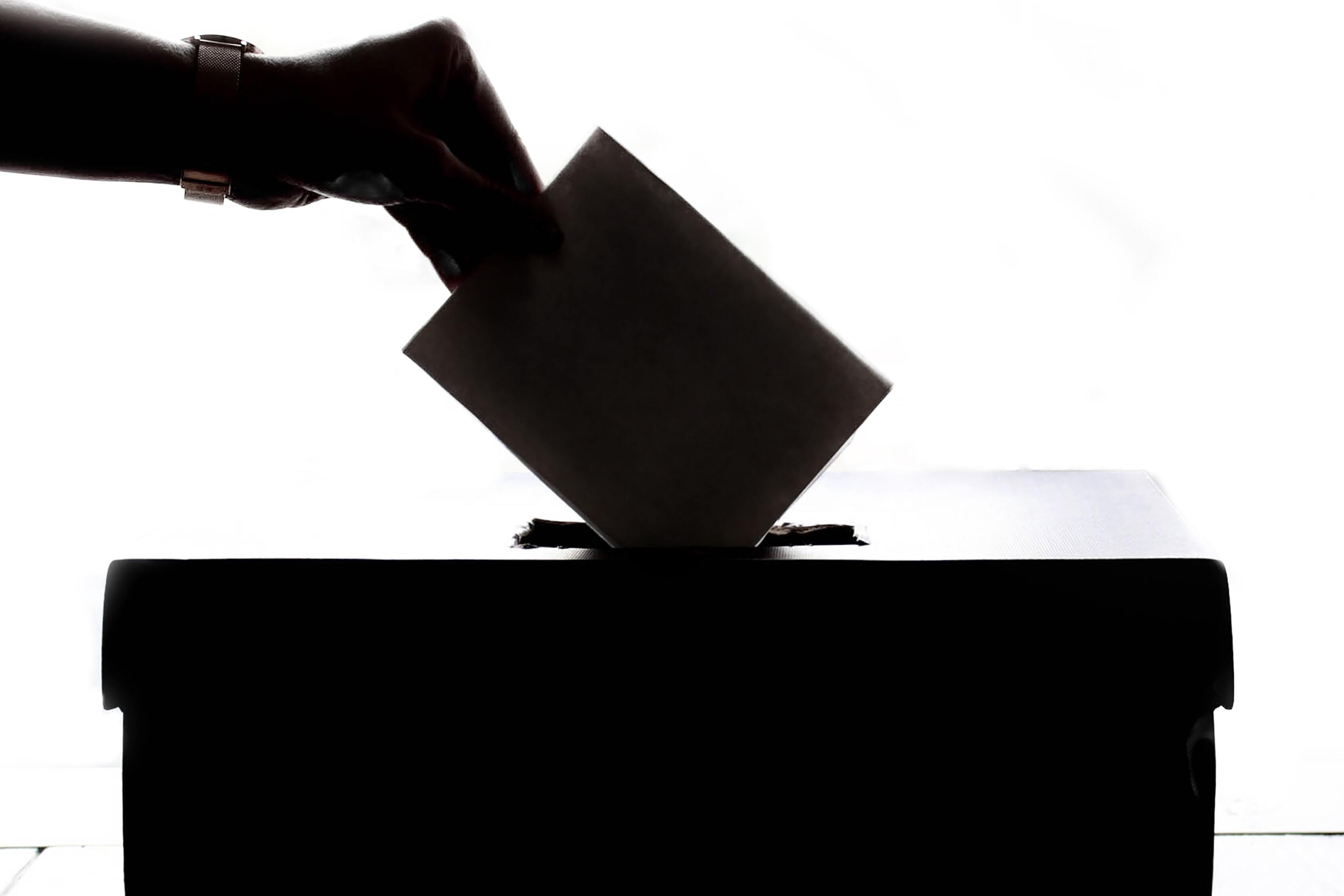 Hand legt Wahlzettel in eine Wahlurne, silhouettiert vor hellem Hintergrund.