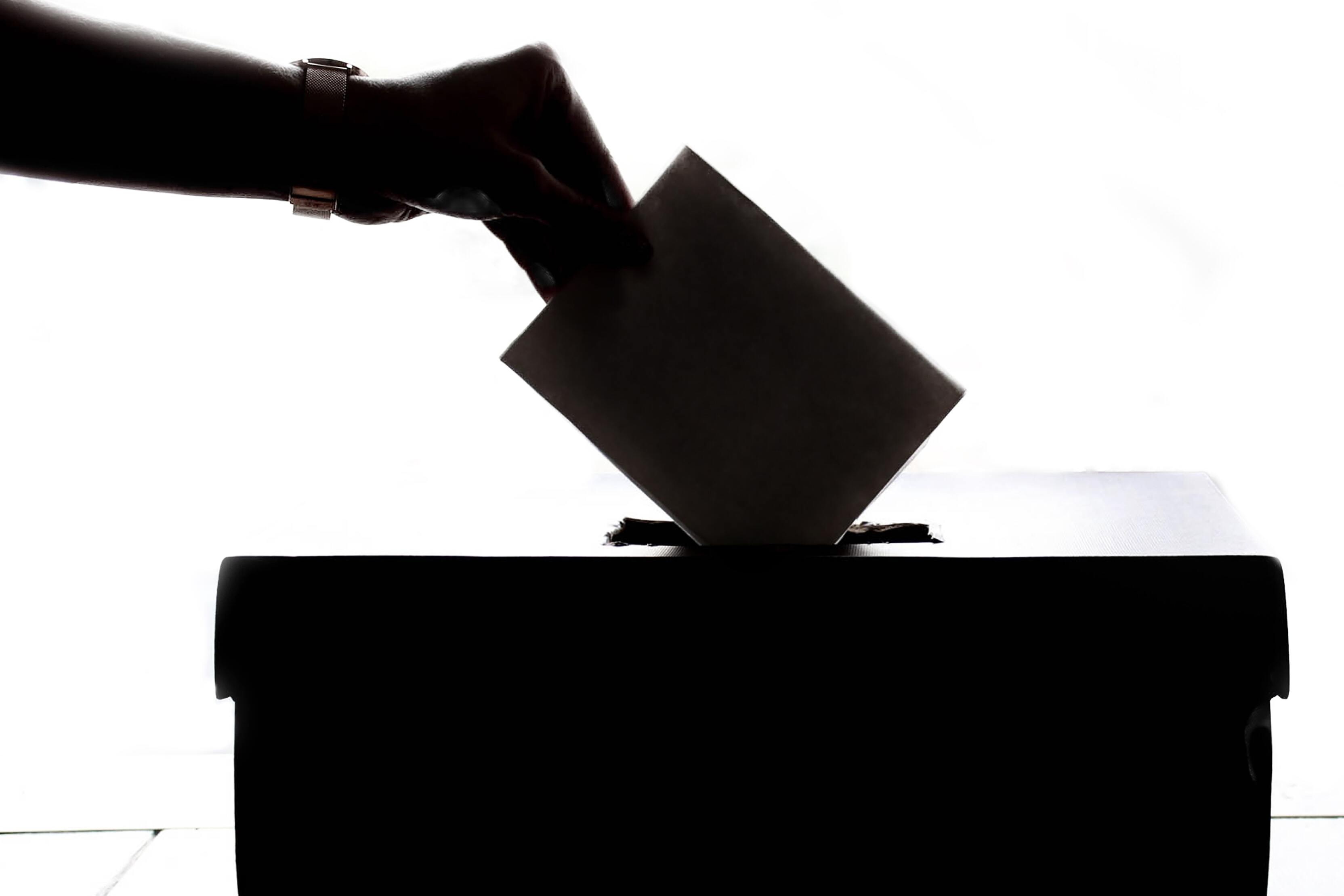 Hand legt Wahlzettel in eine Wahlurne, silhouettiert vor hellem Hintergrund.