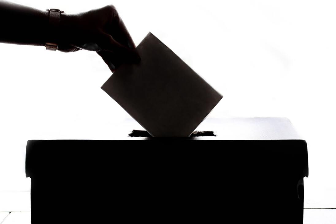 Hand legt Wahlzettel in eine Wahlurne, silhouettiert vor hellem Hintergrund.