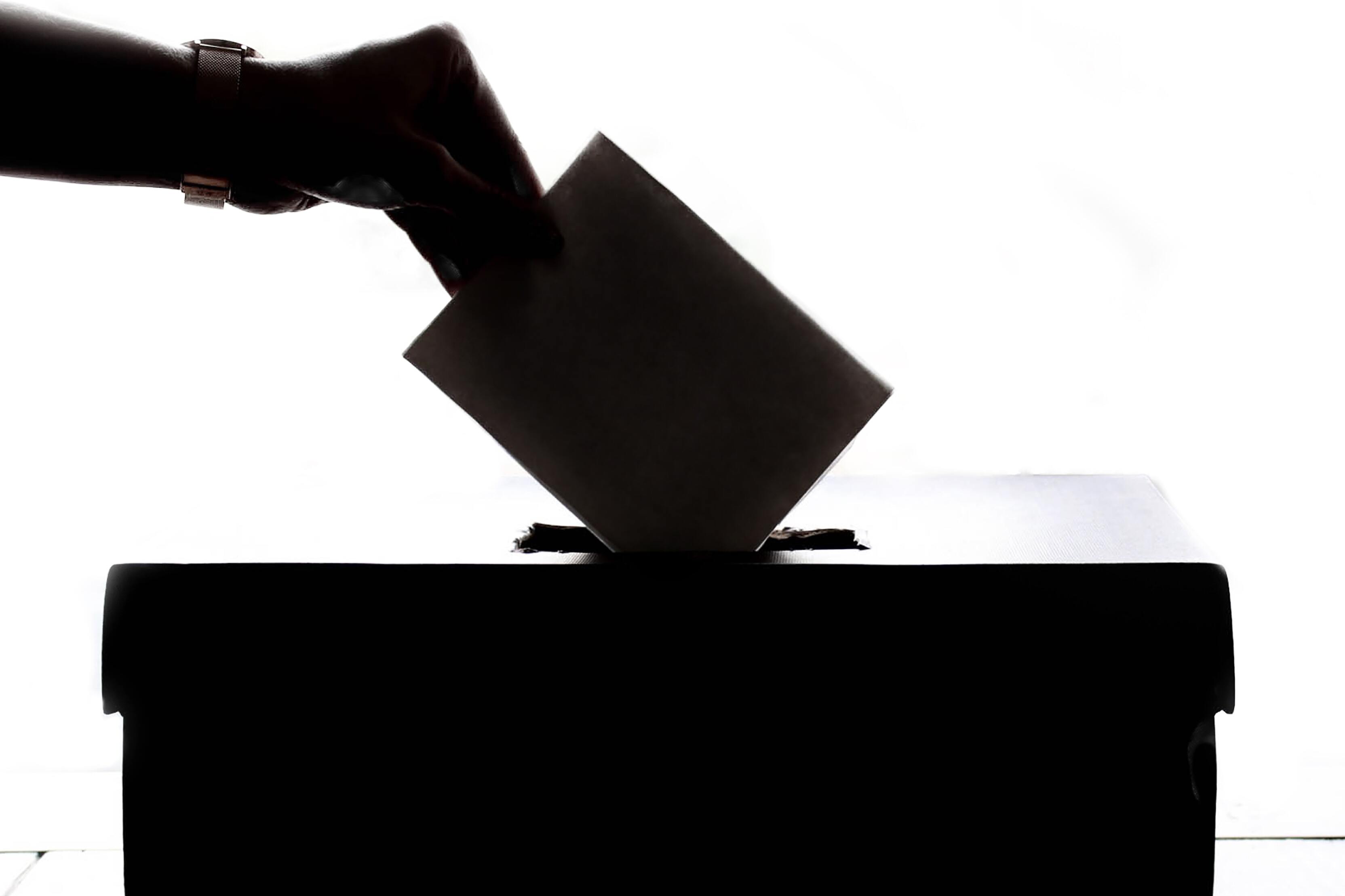 Hand legt Wahlzettel in eine Wahlurne, silhouettiert vor hellem Hintergrund.