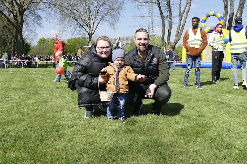 Familie posiert auf einer gr&uuml;nen Wiese: Eltern mit Kleinkind, im Hintergrund Spielger&auml;te und Menschen auf einer Veranstaltung.