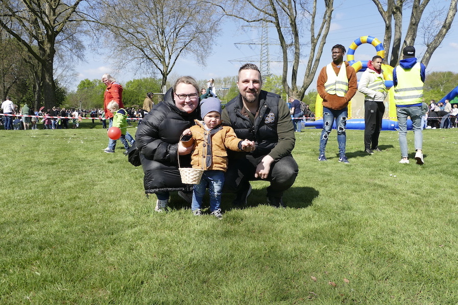 Familie posiert auf einer gr&uuml;nen Wiese: Eltern mit Kleinkind, im Hintergrund Spielger&auml;te und Menschen auf einer Veranstaltung.