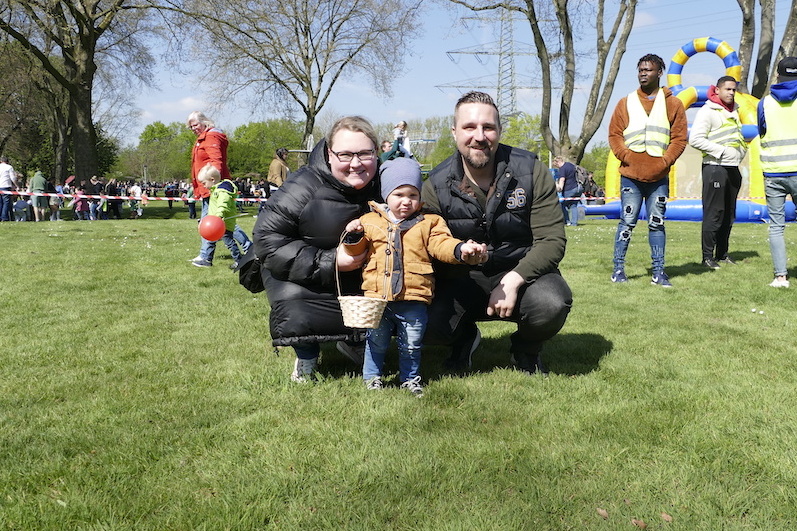 Familie posiert auf einer gr&uuml;nen Wiese: Eltern mit Kleinkind, im Hintergrund Spielger&auml;te und Menschen auf einer Veranstaltung.
