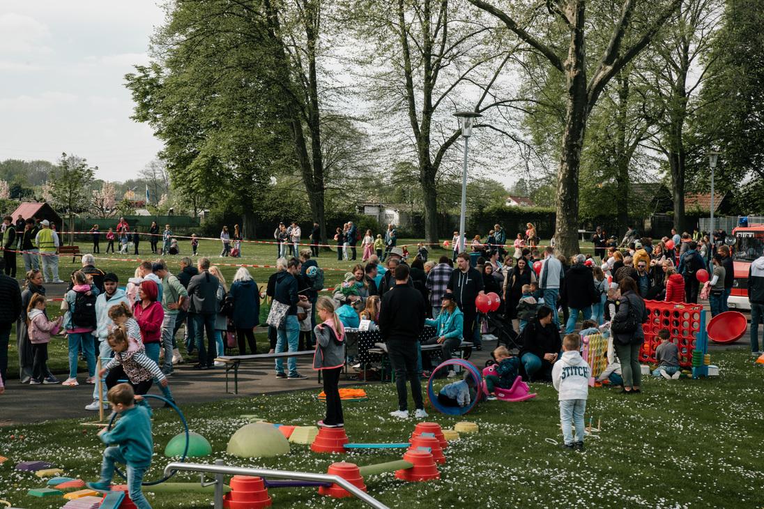 Menschenmenge in einem Park mit Kindern, Spielger&auml;ten und Aktivit&auml;ten unter B&auml;umen an einem sonnigen Tag.