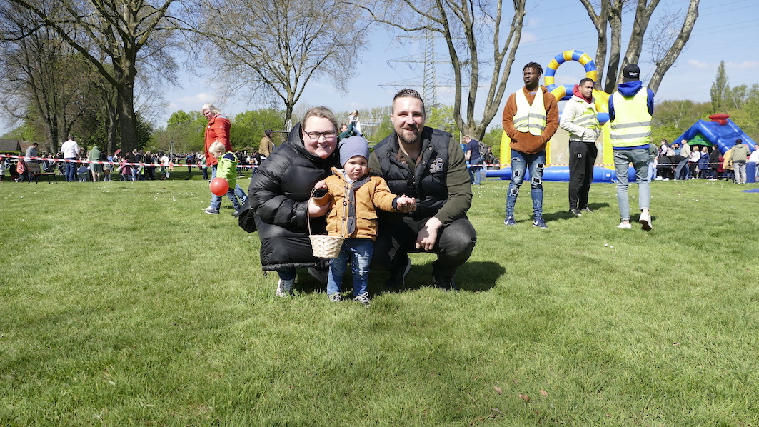 Familie posiert auf einer gr&uuml;nen Wiese: Eltern mit Kleinkind, im Hintergrund Spielger&auml;te und Menschen auf einer Veranstaltung.