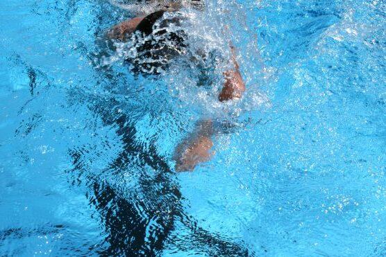 Schwimmer taucht ins Wasser, Arm zieht durch die hellblaue Wasseroberfl&auml;che, Spritzwasser bildet Wellen.