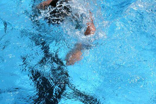 Schwimmer taucht ins Wasser, Arm zieht durch die hellblaue Wasseroberfläche, Spritzwasser bildet Wellen.