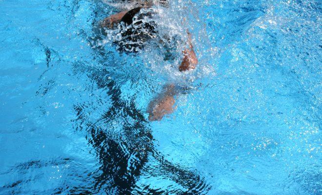 Schwimmer taucht ins Wasser, Arm zieht durch die hellblaue Wasseroberfläche, Spritzwasser bildet Wellen.