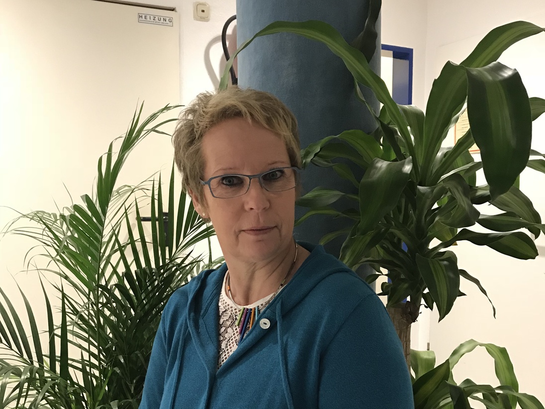 Frau mit Brille und blauer Jacke sitzt vor gr&uuml;nen Pflanzen in einem hellen Raum.