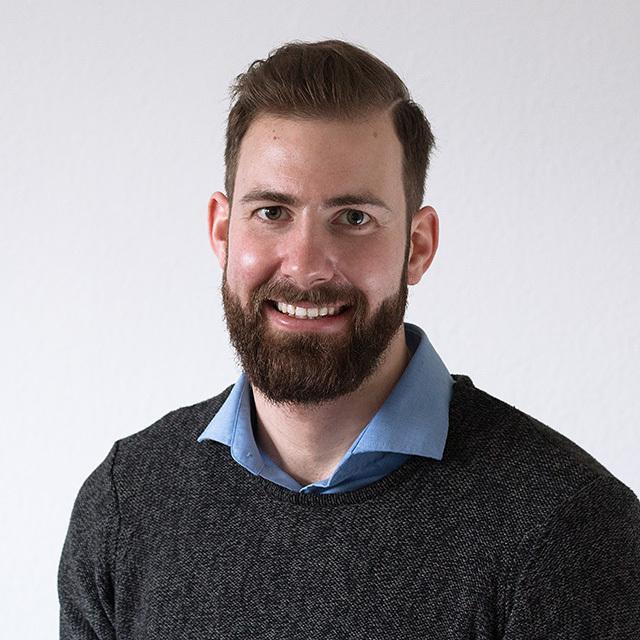 Portrait eines l&auml;chelnden Mannes mit Vollbart, der einen grauen Pullover und ein hellblaues Hemd tr&auml;gt. Helle Wand im Hintergrund.